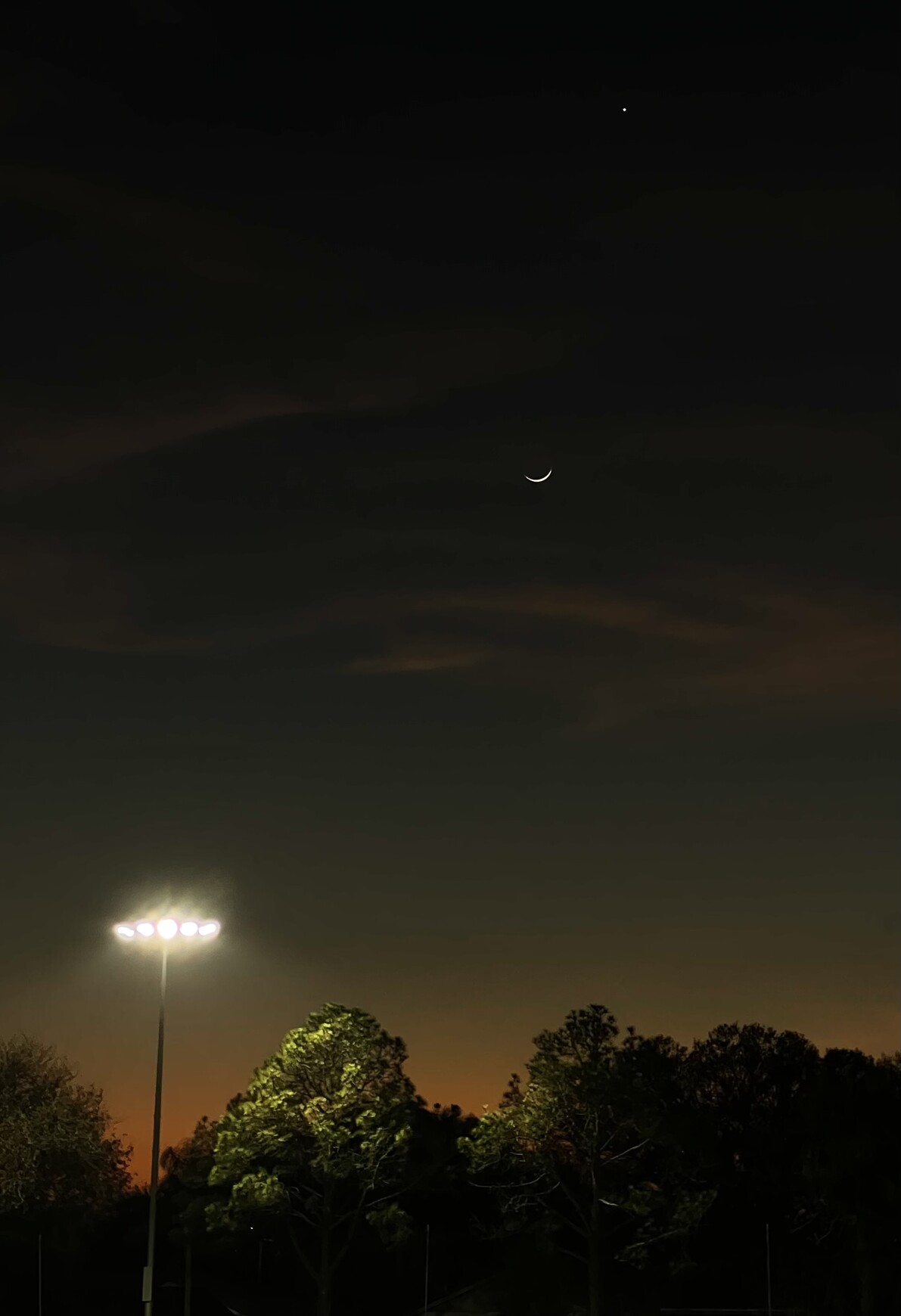 late sunset, orange only low near the horizon, eventually pitch black above. a bright artificial light hangs low over the orange horizon, above which a new moon in the blackness, above that the bright dot of venus.